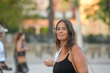 A woman with long hair is smiling and holding a purse. She is surrounded by other people on a sidewalk