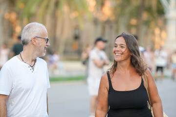 A man and a woman are smiling at each other in a park. The man is wearing a white shirt and the woman is wearing a black tank top