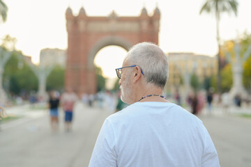 A man wearing glasses and a white shirt stands in front of a large archway. He looks to his left and is surrounded by a crowd of people