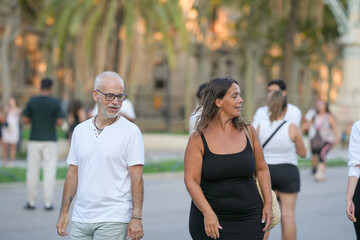 A man and a woman are walking down a street. The man is wearing a white shirt and the woman is wearing a black dress. There are several other people walking around them, some carrying handbags