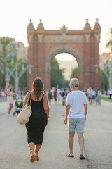 A woman and a man walk down a street in front of a large arch. The woman is carrying a woven bag. The scene is lively and bustling with people walking around