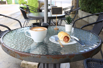 Coffee latte art with sausage roll pastry on glass table in outdoor cafe garden