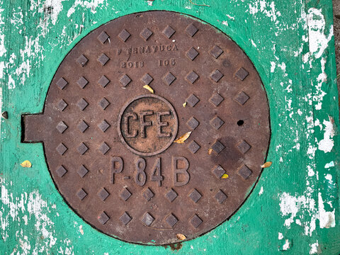 Huatulco, Oaxaca, Mexico. Dec 28, 2025. Weathered Iron CFE Manhole Cover with Embossed Logo on Green Textured Concrete in Mexico