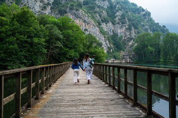A family of three is walking across a wooden bridge over a river. The woman is carrying a child on her back