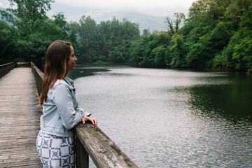 A woman stands on a wooden bridge overlooking a lake. The water is calm and the sky is cloudy. The woman is lost in thought, possibly contemplating the beauty of nature or her own thoughts