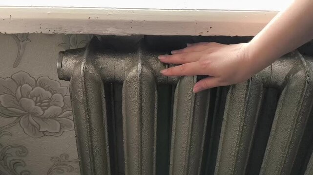 A woman touches an old heating radiator in the house with her hand. Woman warming her hands with old radiator