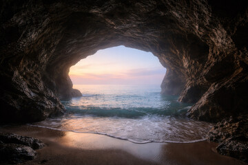 Beautiful Sunrise View From Inside A Dark Coastal Sea Cave Overlooking Gentle Ocean Waves And Sandy Beach