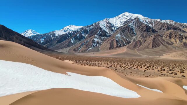 Snow patches on sand dunes with majestic snowcapped mountains under bright blue sky, 4k high quality