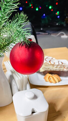 Close-up of a red matte Christmas ball on a spruce branch in a cafe setting with a blurred cake and cookies in the background.