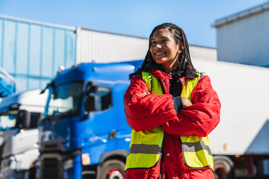 Female professional in reflective vest and warm jacket standing proudly with crossed arms against parked trucks and warehouse