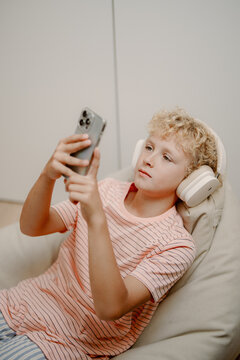 A young boy lounges on a comfortable bean bag chair, focused on his smartphone. He wears headphones and enjoys his time with digital entertainment, looking relaxed.