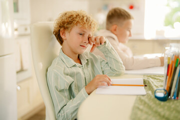 Two children are seated at a table in a bright kitchen. One child looks thoughtful with a notebook in hand, while the other focuses on their work. Sunlight streams in through the window.