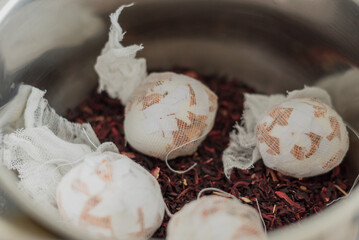 Five eggs wrapped in cloth resting on dried hibiscus petals in a metal bowl. The scene highlights natural dyeing techniques for Easter eggs.
