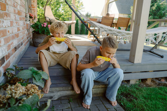 Two boys sit on the porch steps, happily eating fresh corn on the cob. The sun shines down, creating a cheerful atmosphere in a relaxed backyard setting.