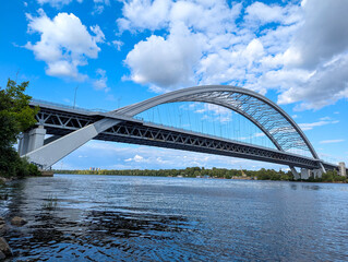 A bridge arch over river. An openwork architectural engineering structure for logistics cargo transportation across body of water. Lower angle shot shore, perspective road. Cumulus clouds landscape