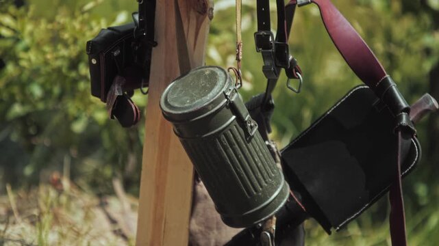 Close-up of German WWII era soldier equipment hanging from wooden post, featuring metal gas mask canister, folded entrenching tool with cover, and camouflage uniform smock.