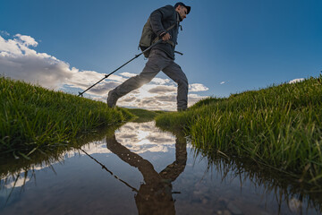 A hiker with poles and a backpack steps across a stream, his reflection visible in the water. Wide side view captures him left to right during golden hour, with lush green banks.