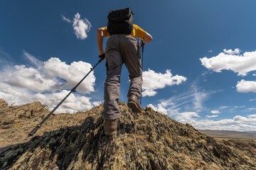 A hiker with backpack and poles ascends a sharp, rocky mountain in the Altai. Medium rear shot from a low angle captures the challenging climb against a blue summer sky with clouds.