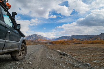 An off-road vehicle is parked on a dirt road overlooking a golden autumn forest and partially snow-capped mountains. A medium shot of its right front tire shows aggressive tread.