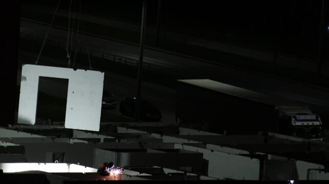 Industrial night scene with worker performing welding task on construction site. Bright orange sparks illuminate concrete foundation elements. Huge precast wall section hangs by chains above ground