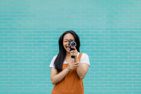 Smiling videographer filming with vintage camera against turquoise brick wall