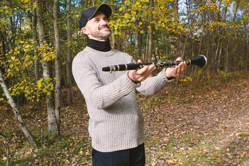 A happy man in a knitted sweater and cap holds a clarinet in an autumn forest, smiling serenely as if listening to nature's melody. Perfect for themes of music, nature, hobbies, and peaceful moments.