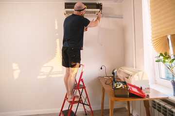 Man on a ladder installing or repairing an air conditioner unit on a white wall. Home improvement, HVAC service, or maintenance work in progress with tools on a nearby table.