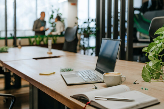 Laptop and coffee cup on modern office desk with two people meeting in background