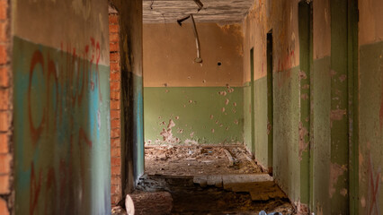 A corridor in an abandoned building shows crumbling walls with peeling paint. Debris is scattered across the floor, and light shines through an opening above.