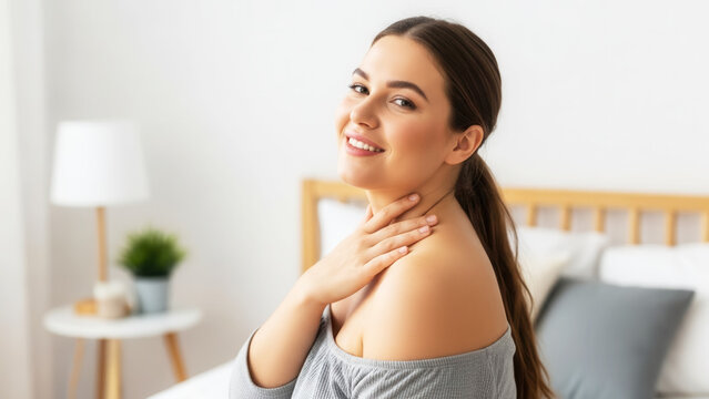 Smiling Caucasian plus size woman bare shoulders in cozy bedroom touching collarbone, body confidence and self care in lifestyle portrait