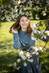 A young Caucasian woman with long brown hair smiles while standing among blooming apple trees. She wears a blue dress and enjoys a sunny day outdoors.