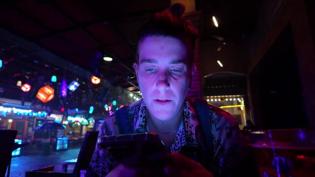 Millennial woman absorbed in her smartphone while sitting in a futuristic looking bar, illuminated by the vibrant, colorful glow of neon lights and her phone's screen in the evening