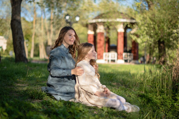 Two young girls sit on the grass in a park. One girl has long brown hair and wears a blue dress. The other girl has curly red hair and wears a light sweater. A gazebo is in the background.