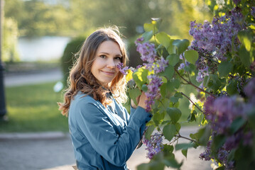 A young Caucasian woman with long brown hair smiles while touching purple lilac flowers in a sunny park setting.