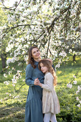 A young Caucasian woman with brown hair and a girl with red hair stand together under a blooming tree in a sunny park. They are smiling and holding hands.
