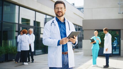 Confident Caucasian male doctor standing outside hospital. Medical specialist holding tablet. Background colleagues discussing cases. Outdoor clinic area hosting professional team.