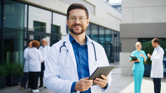 Confident Caucasian male doctor standing outside hospital. Medical specialist holding tablet. Background colleagues discussing cases. Outdoor clinic area hosting professional team. - Powered by Adobe