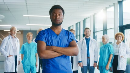 Obraz premium Confident male medical worker crossing arms while standing in bright corridor. Focused nurse presenting strength while group of doctors supporting him in background. Preparing for challenging duties.