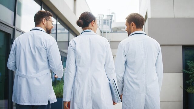 Focused female doctor holding folders and walking between two male colleagues. Confident team moving together and discussing upcoming duties. Professionals continuing route toward clinic entrance. - Powered by Adobe