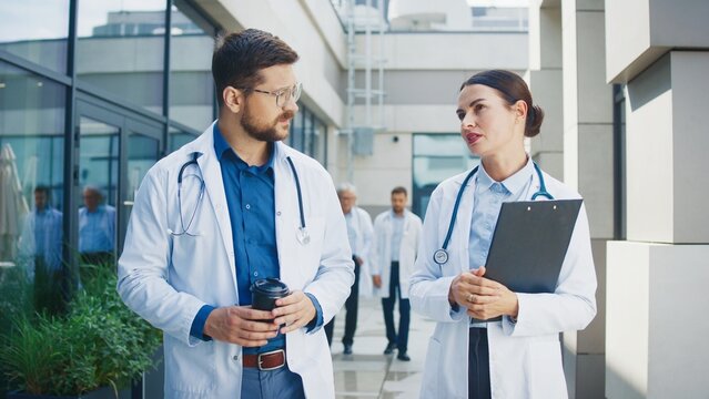 Two doctors walking at hospital complex corridor. Having conversation during break. Male carrying coffee cup while female colleague holding clipboard. Medical staff behind continuing daily tasks. - Powered by Adobe
