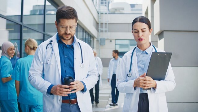 Two doctors walking at hospital complex corridor. Having conversation during break. Male carrying coffee cup while female colleague holding clipboard. Medical staff behind continuing daily tasks. - Powered by Adobe