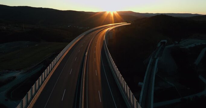 Drone shot flying over a highway bridge at sunset, catching warm reflections, then smoothly moving under the bridge. Calm, cinematic atmosphere with modern infrastructure in Slovenia. Čni Kal Viaduct.