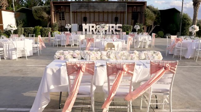 Tables are arranged outside for a wedding celebration. White cloth covers the tables. Pink sashes are tied on chairs. Flowers are placed on tables and decor is set around the area.