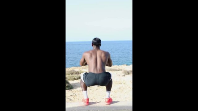 Man doing squats outdoors by the sea. Vertical video of an athletic man performing squats during outdoor fitness training with the sea in the background, promoting strength and healthy lifestyle.