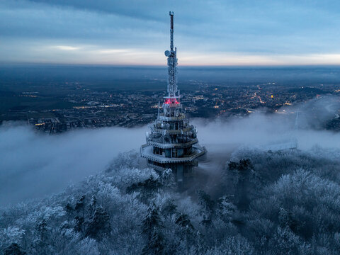 Aerial view of the iconic Zobor transmitter piercing through a sea of fog, its red lights glowing against the twilight sky, above snow-laden trees, Nitra, Nitra Region, Slovakia.