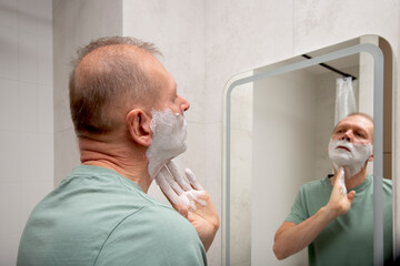 A man applies shaving foam to his neck while looking at his reflection in a bathroom mirror at home.