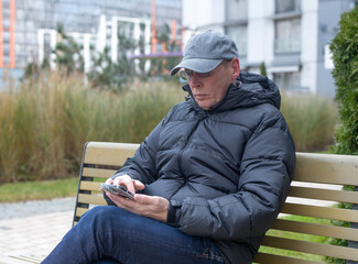 Obraz premium A mature man sits on a bench outdoors while using a smartphone in a residential area. Everyday urban