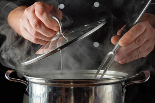 In a busy kitchen, a cook lifts the lid of a pot with his hand and stirs food with a fork. Steam escapes from the pot as the cook inspects the contents, preparing the dish for the customers. - Powered by Adobe