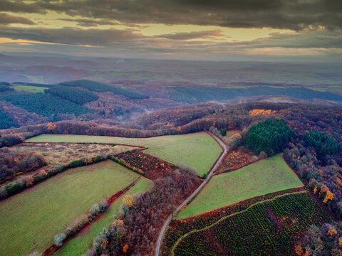 Aerial view of patchwork fields nestled amidst the autumnal tapestry of Morvan's forests, where verdant pastures meet the fiery hues of turning leaves under a brooding sky, Morvan, Morvan, France.