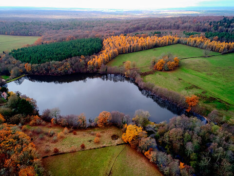 Aerial view of the dark lake reflecting the sky, surrounded by the vibrant autumn colours of the Morvan forest, France.